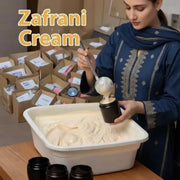 A woman in traditional attire carefully scooping handmade Zafrani Cream into black cosmetic jars from a large container, with packed shipping boxes in the background.